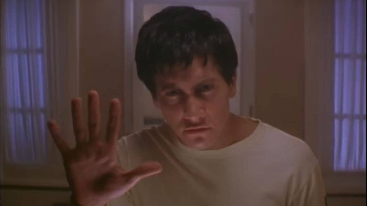 Jake Gyllenhaal looks into a mirror, raising his hand to touch the glass, looking concerned.